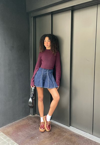 Mujer con cabello rizado que lleva un suéter burdeos, falda azul, calcetines blancos y zapatos marrones, sosteniendo un bolso negro, de pie junto a una pared gris.