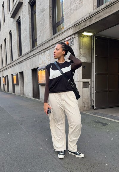 Top sin mangas blanco y negro sobre una camiseta de manga larga marrón, pantalones sueltos beige y zapatillas negras. Sostiene un teléfono en la mano izquierda.