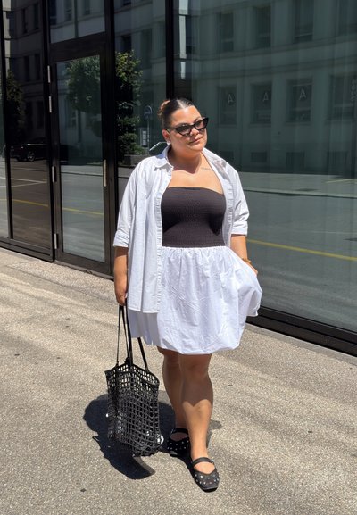 Camisa blanca de botones sobre un vestido negro sin tirantes, con una falda blanca acampanada. Bolso de red negro, zapatos negros con lunares.