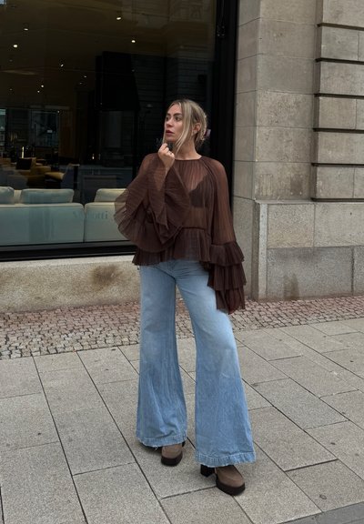 Top marrón con plisados y mangas con volantes, combinado con unos vaqueros de mezclilla claros de tiro alto y de campana. Botas de plataforma marrones y gruesas completan el conjunto.