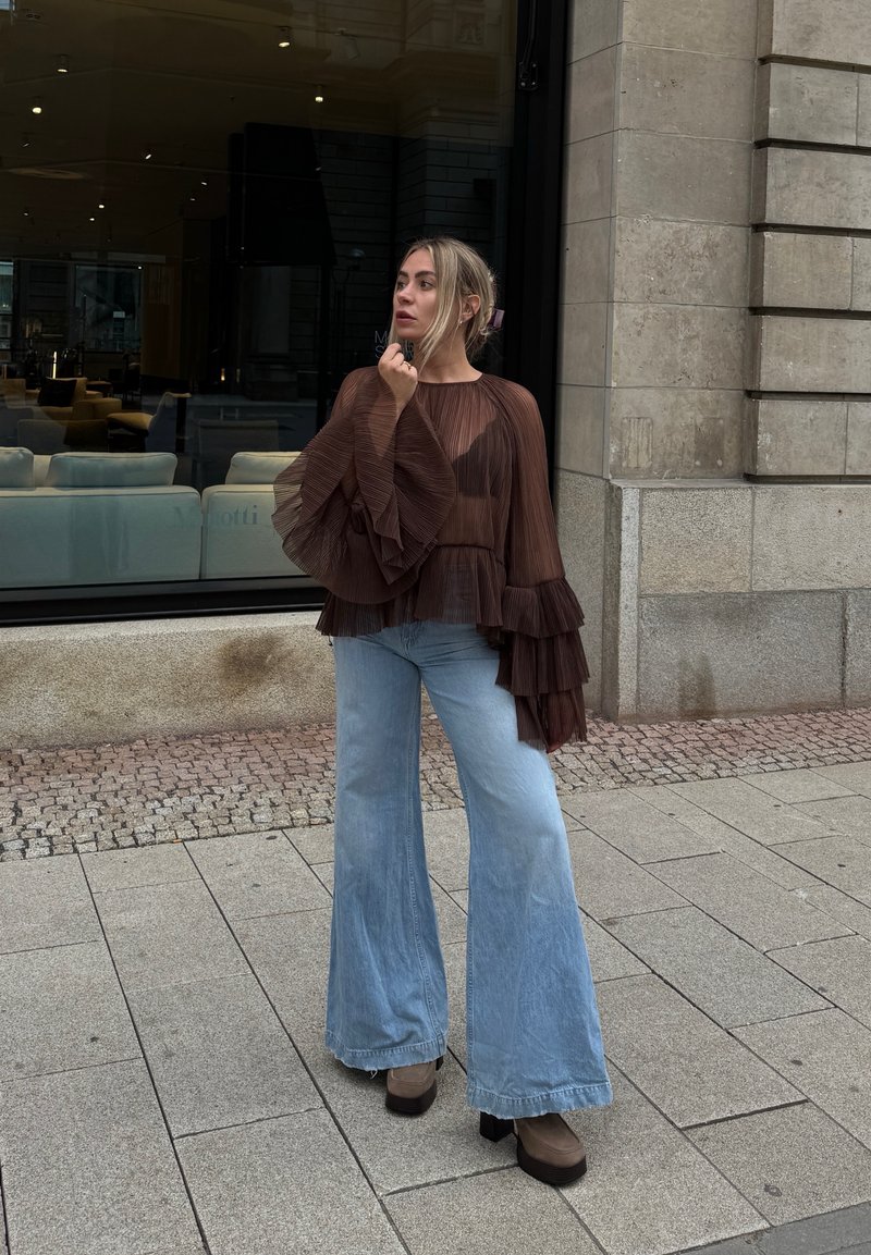 Top castanho plissado com mangas em babados, combinado com jeans flare de cintura alta em denim claro. Botas platform castanhas robustas completam o look.