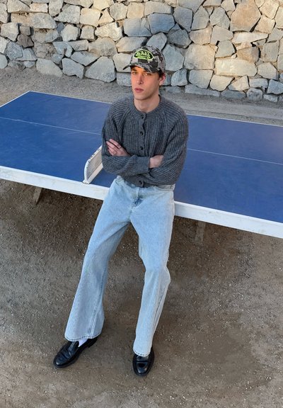 Camisa de manga larga gris con rayas y botones, vaqueros de pierna ancha azul claro y zapatos negros. Gorra de béisbol con camuflaje y un logo.