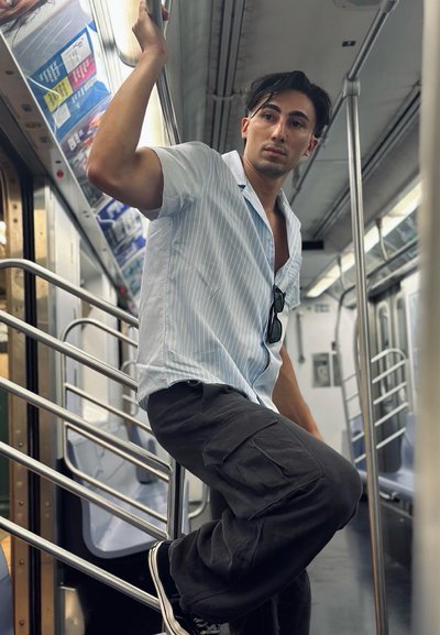 Una persona se apoya en un poste metálico en un vagón de metro, vistiendo una camisa de manga corta a rayas azul claro y pantalones cargo negros.