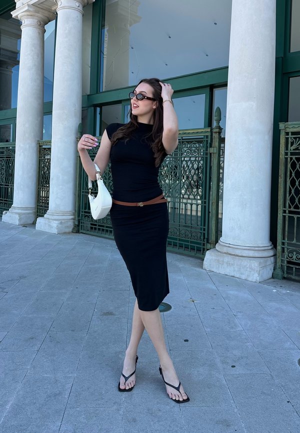 Vestido preto justo, cinto na cintura feito de couro castanho. Usando chinelos pretos, segurando uma mala branca. Ambiente ao ar livre com detalhes verdes.