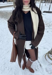 Femme debout sur la neige vêtue d'un manteau marron, d'une écharpe blanche, de gants noirs, d'un pull bleu marine, d'une jupe blanche, de collants noirs et de bottes marron, tenant un sac marron.