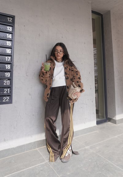 Joven con chaqueta de estampado de leopardo y pantalones holgados, sosteniendo una bebida verde y un bolso beige, de pie frente a una pared gris texturizada cerca de buzones.