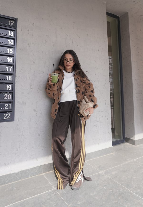 Mlajša ženska, oblečena v jakno z vzorcem leoparda in široke hlače, drži zeleno pijačo in bež torbico, stoji ob teksturiranem sivem zidu blizu poštnih nabiralnikov.