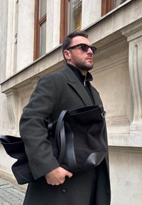 Homme portant un manteau sombre et des lunettes de soleil, portant un grand sac en cuir et tissu noir, debout à côté d'un bâtiment beige avec de grandes fenêtres.