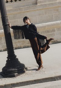 Jeune femme en haut noir à franges et pantalon marron, tenant un lampadaire, levant une jambe derrière elle sur un trottoir en ville avec un mur en pierre en arrière-plan.