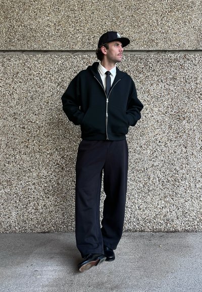 Sudadera negra con cremallera sobre una camisa blanca de cuello y corbata negra, combinada con pantalones holgados negros y zapatos negros. El fondo es de hormigón texturizado.