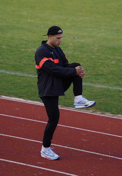 Hombre atlético con chaqueta negra con rayas naranjas y gorra negra, estirándose en una pista de atletismo, vistiendo zapatillas deportivas blancas y azules.