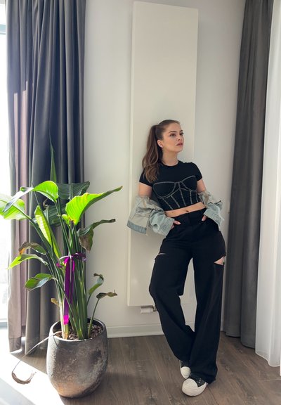 Top corto negro con embellishments plateados, chaqueta de mezclilla clara, pantalones anchos negros con cortes y zapatillas blancas. Planta verde en maceta en primer plano.