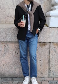 Sort blazer over en hvid T-shirt, let brun sweater bundet omkring halsen, blå jeans og hvide sneakers. Holder en kaffekop.