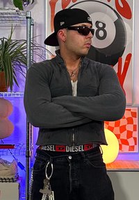 Μαύρο κοντό ζιπ-ζακέτα, σκούρα τζιν, γυαλιά ηλίου και καπέλο. Έντονη ζώνη Diesel, μπρελόκ και επίπεδες αλυσίδες. Διακοσμητικό φόντο.