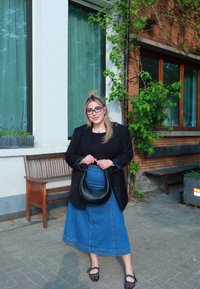 Czarna marynarka, czarny top i niebieska spódnica dżinsowa. Czarna torebka o zakrzywionym kształcie. Sandały z koralikami. Zielone tło z drewnianą ławką.