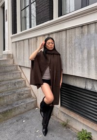 Poncho en tricot marron avec une capuche, superposé à un haut gris et un short noir, associé à des bottes en fourrure noires longues et des escarpins noirs pointus.