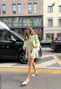 Manteau vert en peluche, blouse beige clair, jupe argentée et sac à main blanc. Le mannequin porte des mules blanches et marche dans une rue de la ville près de voitures garées.