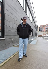 Homme portant une casquette noire, une veste zippée noire, un jean bleu clair et des baskets noires, debout sur un trottoir urbain près d'un grand immeuble.