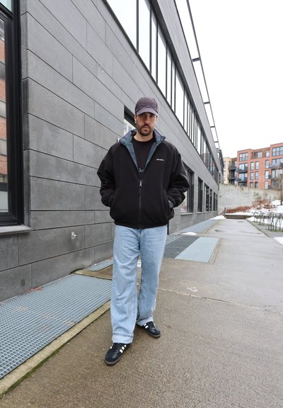 Hombre con gorra negra, chaqueta negra con cremallera, vaqueros azul claro y zapatillas negras de pie en la acera urbana cerca de un edificio alto.