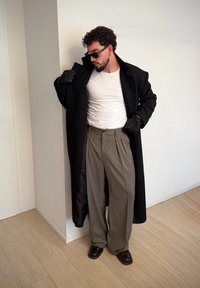 Long manteau noir, chemise blanche, pantalon gris taille haute, gants en cuir noir et chaussures brun foncé. Textures lisses avec une coupe ajustée.