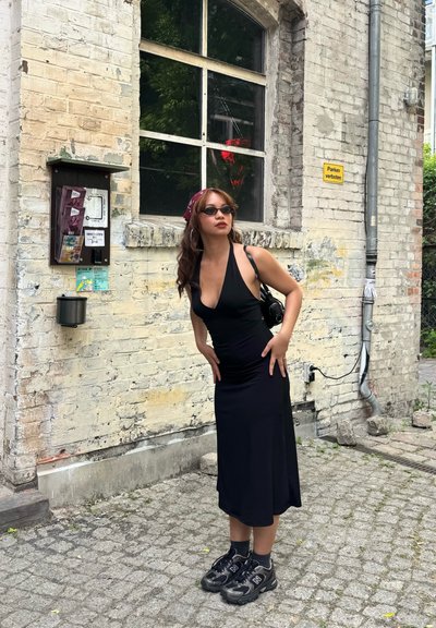 Joven mujer con vestido negro y zapatillas posa frente a una pared de ladrillo envejecida con ventana, carteles y un letrero que dice “Parken verboten”.