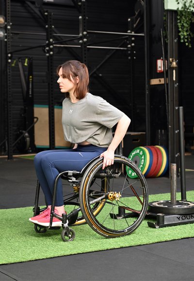 Mujer en silla de ruedas con ropa deportiva, preparándose para hacer ejercicio en un gimnasio con discos de pesas y equipo cerca.