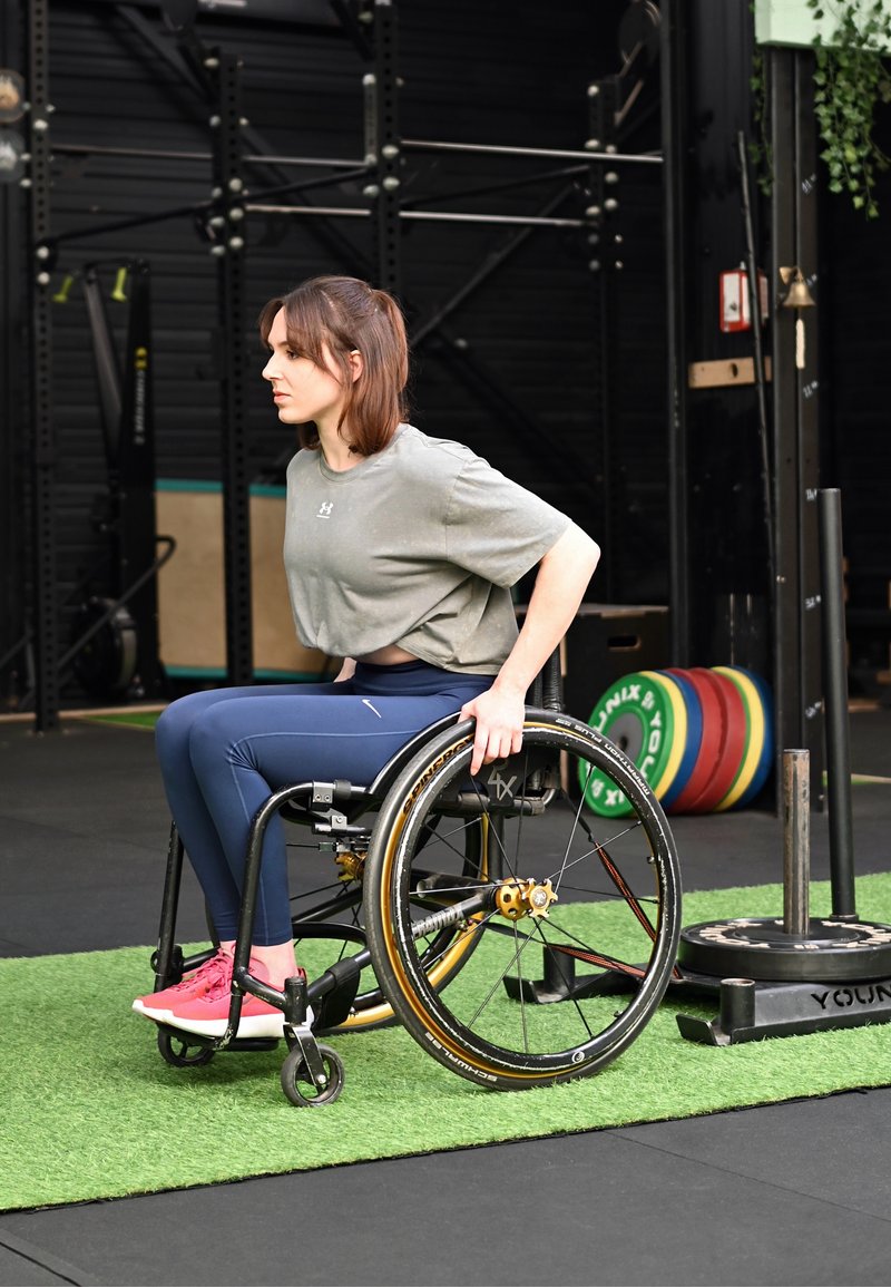 Femme en fauteuil roulant portant des vêtements de sport, se préparant à faire de l'exercice dans une salle de sport avec des disques de poids et du matériel à proximité.
