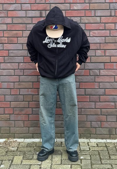 Sudadera negra con cremallera con "Low Lights Studios" en letras blancas, jeans rectos azul claro y zapatos de plataforma negros, dando la espalda a una pared de ladrillos.