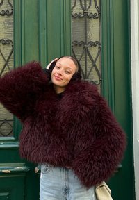 Burgundska umetna krzna jakna z oversized rokavi, teksturirana površina, v kombinaciji s svetloblue denim hlačami in črnimi ušesnimi zaščitniki, postavljena pred zelenimi vrati.