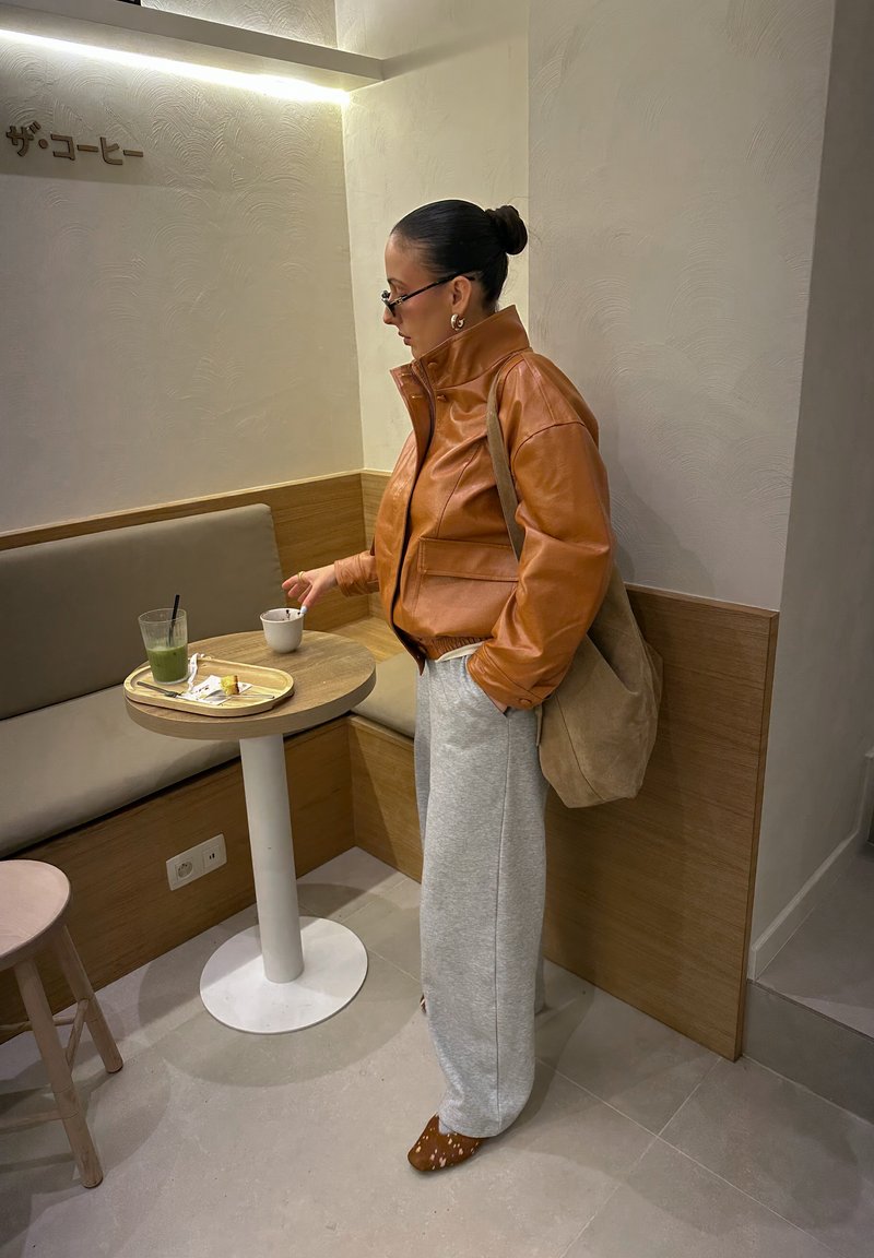Veste en cuir marron, pantalon de survêtement gris et chaussures marron avec un motif. Debout à côté d'une table ronde avec des boissons et de la nourriture sur un plateau.