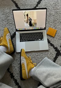 Κίτρινα σουέτ αθλητικά παπούτσια με λευκές λεπτομέρειες, τοποθετημένα σε μια υφασμάτινη γκρίζα μοκέτα, δίπλα σε έναν φορητό υπολογιστή και μια κίτρινη θήκη κάρτας.