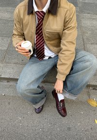 Pruun krae jakki valge särk, punaste triipudega lips, heledad sinised teksad, mille säärtel on üles keeratud, ja burgundi punased mokassiinid. Käes tass.