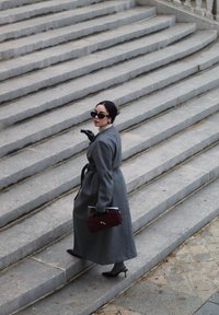 Grauer Wollmantel mit Gürtel, langen Ärmeln und hohem Kragen. Schwarze Handschuhe, runde Sonnenbrille und eine burgunderfarbene Handtasche. Steinige Treppe im Hintergrund.