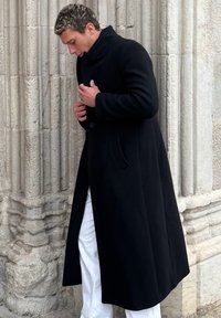 Homme ajustant un manteau long noir à col montant, debout près d'un mur en pierre texturé, portant un pantalon blanc.