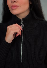 Μαύρος πλεκτός σκούφος με ψηλό γιακά και ασημί φερμουάρ. Το μοντέλο κρατά το φερμουάρ με το ένα χέρι, επιδεικνύοντας μακριά, περιποιημένα νύχια.