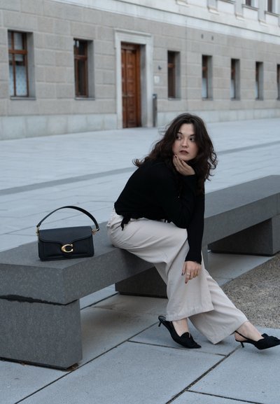 Top negro de manga larga, pantalones beige de pierna ancha, bolso pequeño negro con herrajes dorados, y zapatos de tacón negros sobre un banco de piedra.