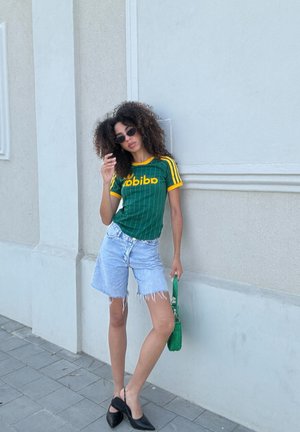 Camiseta a rayas verde y amarilla con el texto "babibo", pantalones cortos de mezclilla desgastados, tacones negros con punta y un bolso de mano tejido verde.