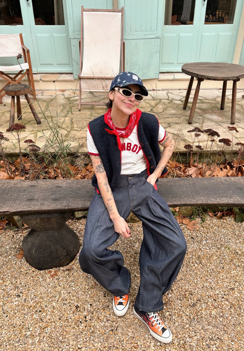 Personne portant une casquette à motifs d'étoiles, un bandana rouge, un t-shirt blanc "COWBOY", un gilet noir, un pantalon large et des baskets à motifs de flammes, assise sur un banc en bois en plein air.