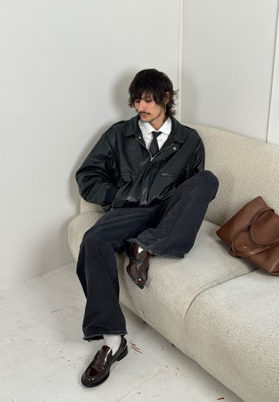 Chaqueta bomber de cuero negro, camisa blanca, corbata negra, jeans oscuros, zapatos marrones y un bolso de cuero marrón sobre un sofá texturizado color crema.