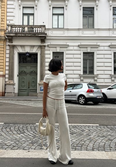 Top de punto blanco con estampado de estrellas, pantalones blancos con cordón, y un pequeño bolso blanco; zapatos negros; fondo de calle de adoquines.