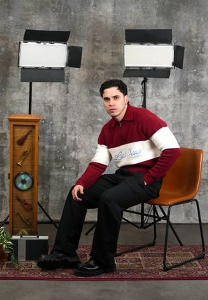 Hombre con suéter rojo y blanco sentado en una silla marrón junto a un armario con temática musical, una radio vintage y dos luces de estudio sobre un fondo gris.