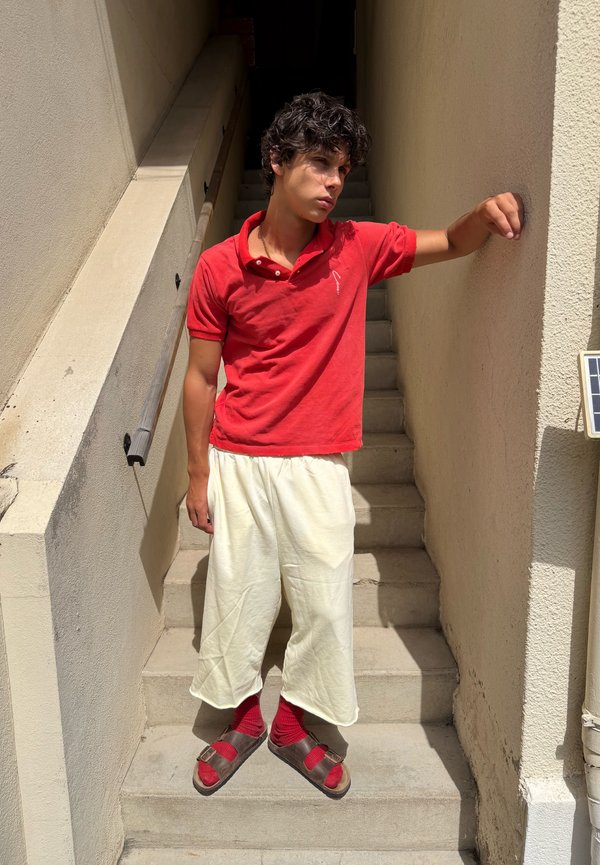 Jeune homme en chemise rouge et pantalon blanc debout sur des escaliers extérieurs, appuyé d’un bras contre un mur beige, regardant pensivement sur le côté.