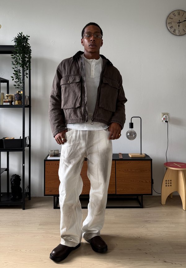 Un hombre posa con una chaqueta marrón oscuro, camisa y pantalones blancos y zapatos negros en una habitación amueblada.