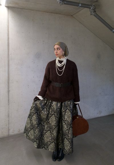 Mujer con un suéter marrón, pañuelo de cabeza de color oliva, collares de perlas en capas, falda larga estampada, sosteniendo un bolso marrón contra una pared de cemento.
