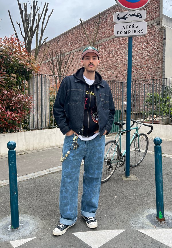 Hombre con chaqueta vaquera, vaqueros estampados y gorra, de pie con las manos en los bolsillos cerca de una bicicleta y un cartel que dice “Accès Pompiers” en la calle.