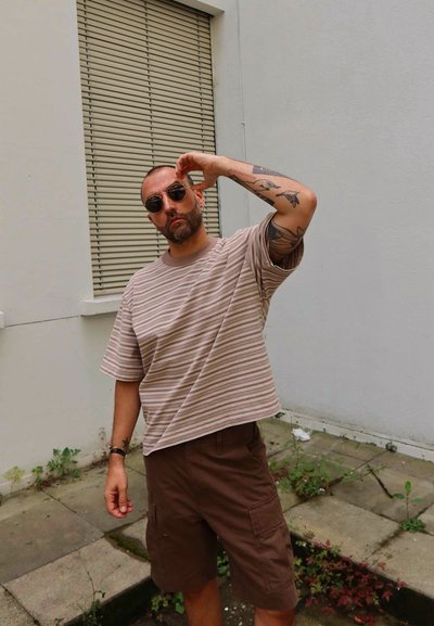 Camiseta extra grande a rayas en beige y marrón con cuello redondo, combinada con pantalones cortos cargo marrones. Los accesorios incluyen gafas de sol redondas oscuras.