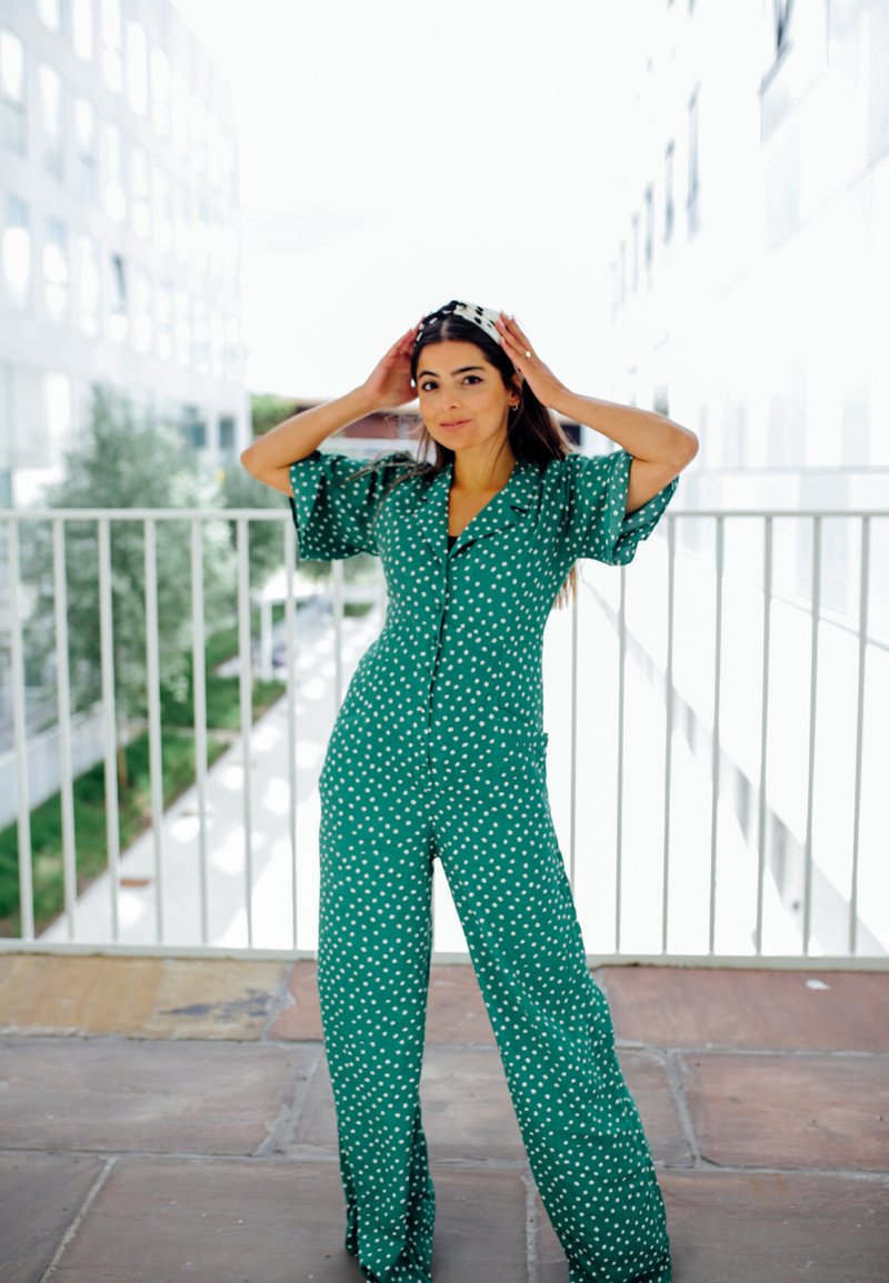 Zöld pöttyös overall rövid ujjal, gombos záródással és bő szárakkal. Az anyag sima textúrájú, világos háttér előtt.