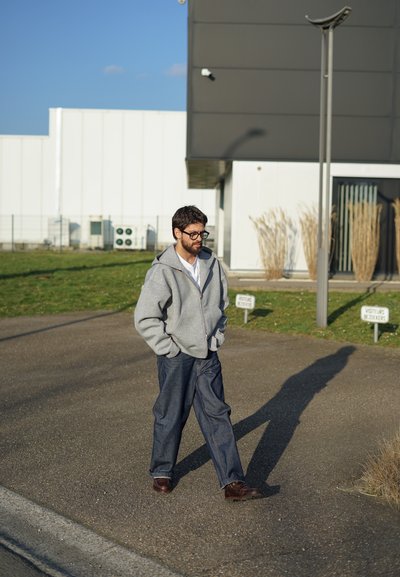 Sudadera gris con cremallera, camiseta blanca, jeans oscuros holgados y zapatos marrones. Caminando al aire libre con un fondo de edificios y hierba.
