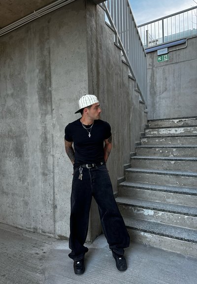 Camiseta negra, pantalones de mezclilla negros holgados, zapatillas negras y una gorra blanca con rayas negras. Los accesorios incluyen un collar y un llavero.