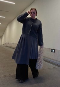 Grauer, strukturierter Cardigan mit runden Knöpfen über einem gefalteten dunklen Rock. Schwarze, weit geschnittene Hosen und ein flauschiges, helles Accessoire. Urbane Umgebung.